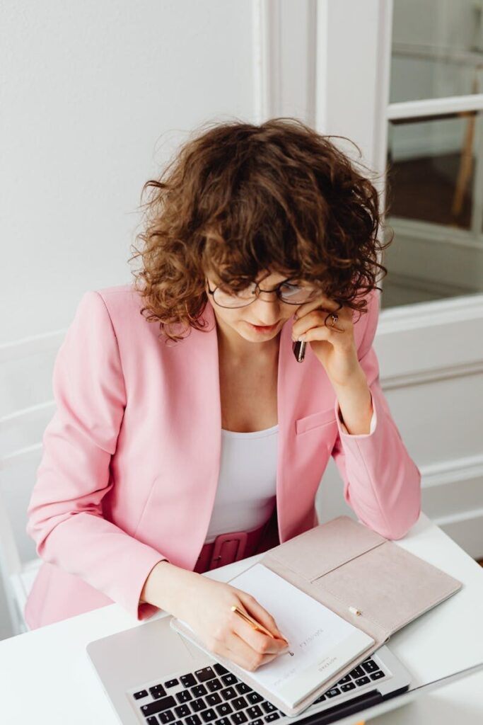 Caucasian businesswoman multitasking at her desk with a phone and notebook in a modern office.